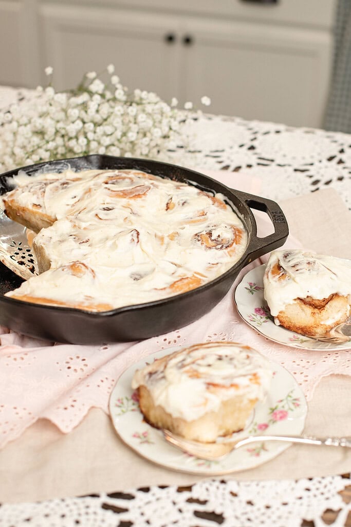 Cast iron skillet of sourdough cinnamon rolls generously frosted with cream cheese frosting beside two individual rolls served on floral china plates, with baby's breath in the background on a lace doily