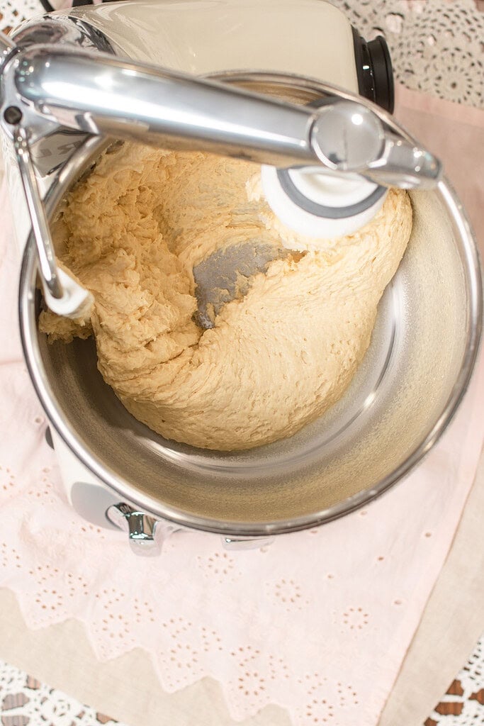 Close-up overhead view of sourdough cinnamon roll dough being kneaded in a stand mixer bowl with a dough hook, on a pink eyelet linen