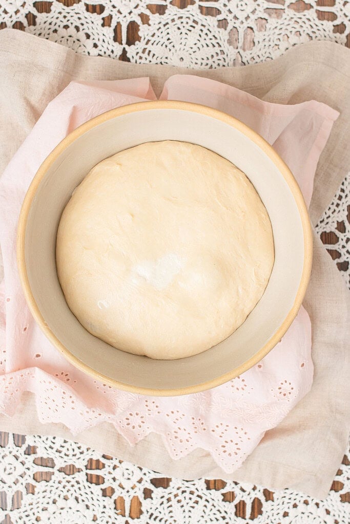 Overhead view of sourdough cinnamon roll dough fully risen in a smooth dome in a ceramic proofing bowl, on a pink eyelet linen and lace doily