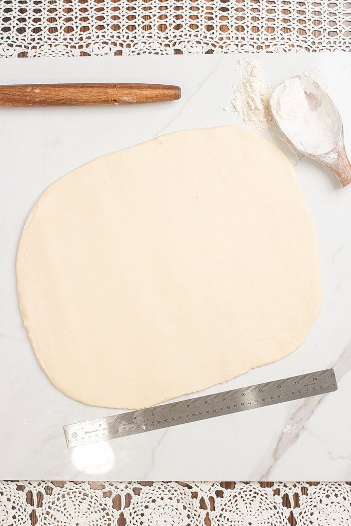 Overhead view of sourdough cinnamon roll dough rolled into a flat rectangle on a marble surface, with a wooden rolling pin, a flour scoop, and a metal ruler alongside