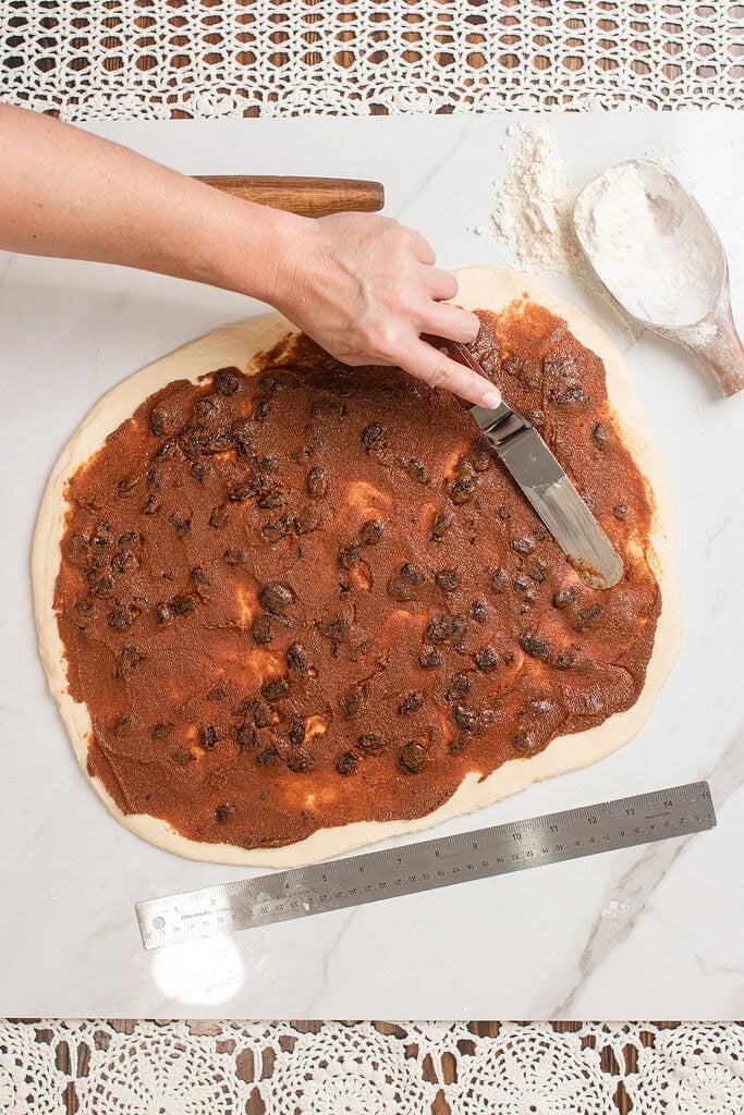 Hand spreading cinnamon brown sugar butter filling across rolled-out sourdough cinnamon roll dough with an offset spatula on a marble surface, with a flour scoop and metal ruler alongside