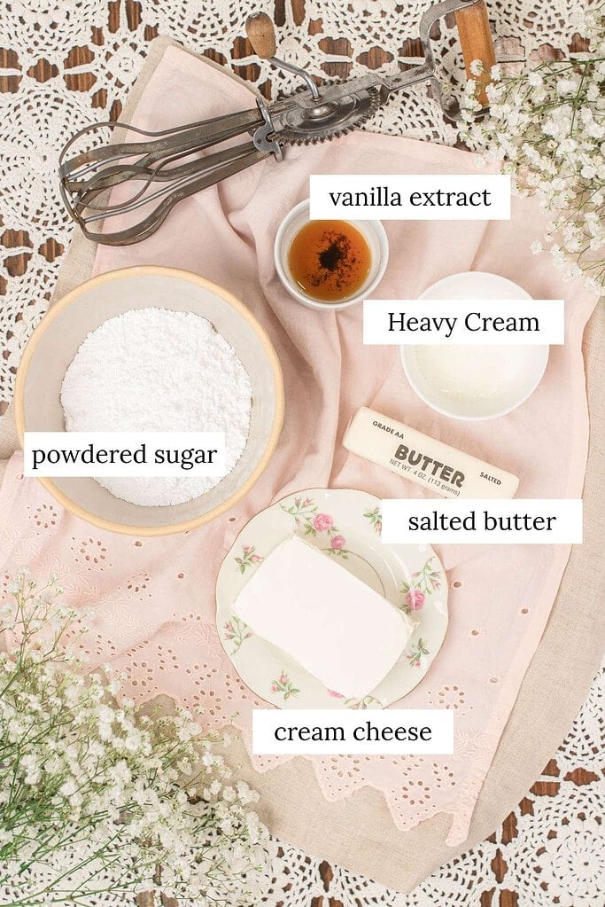Flat lay of cream cheese frosting ingredients on a pink eyelet linen -- a bowl of powdered sugar, a block of cream cheese on a floral china plate, a stick of salted butter, a small bowl of vanilla extract, a small bowl of cream, and a vintage hand mixer