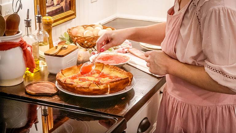 a women in a cottage kitchen drizzling strawberry compote on a einkorn dutch baby pancake
