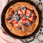 A puffed Dutch baby pancake in a black cast iron skillet topped with fresh strawberries and blackberries and lightly dusted with powdered sugar.
