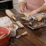 Two women are rolling pasta sheets through a manual pasta roller in a kitchen on a wooden counter top.