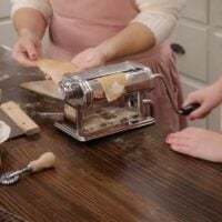 Two women are rolling pasta sheets through a manual pasta roller in a kitchen on a wooden counter top.