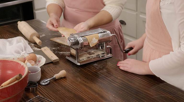 Two women are rolling pasta sheets through a manual pasta roller in a kitchen on a wooden counter top.