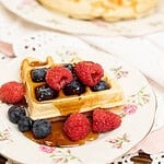 Sourdough discard waffle topped with raspberries, blueberries, and maple syrup served on a vintage floral plate with another waffle in the background.