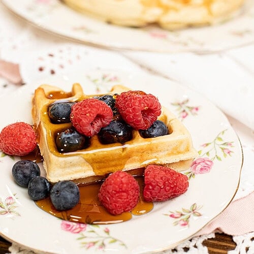 Sourdough discard waffle topped with raspberries, blueberries, and maple syrup served on a vintage floral plate with another waffle in the background.