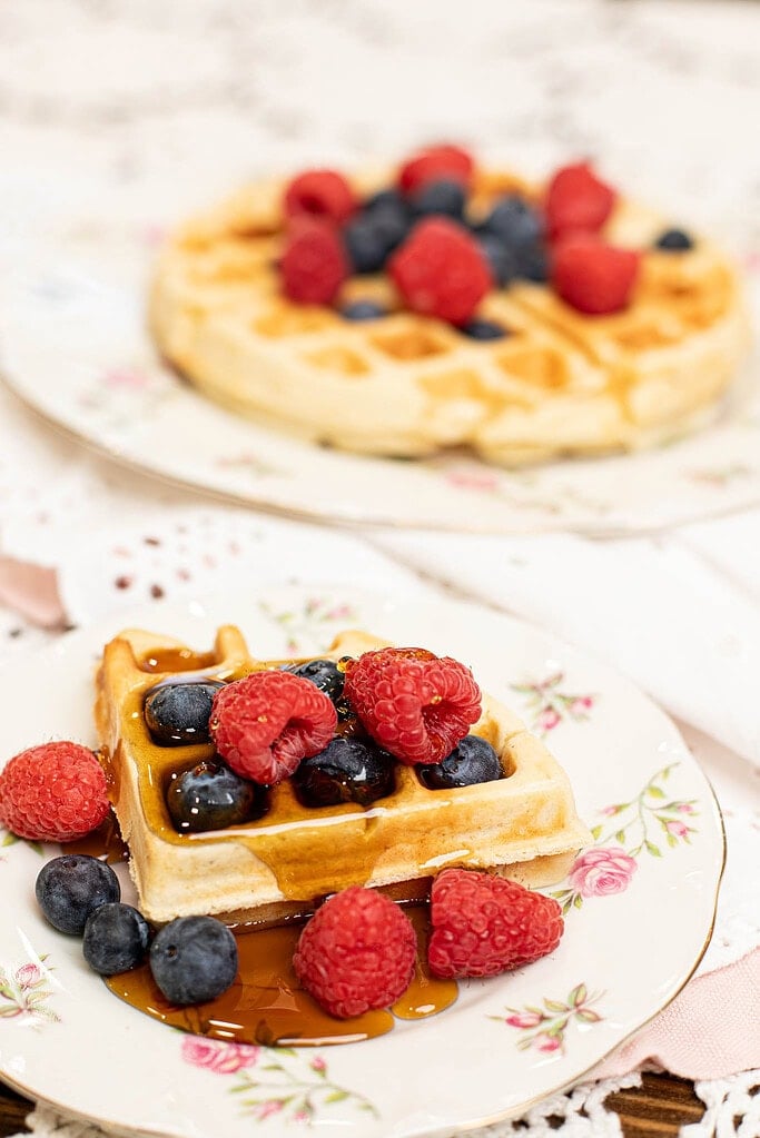 Sourdough discard waffle topped with raspberries, blueberries, and maple syrup served on a vintage floral plate with another waffle in the background.