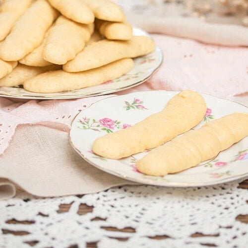 Two floral china plates of sourdough ladyfingers on a pink linen -- a large plate piled high and a small plate with two ladyfingers -- with dried baby's breath in the background