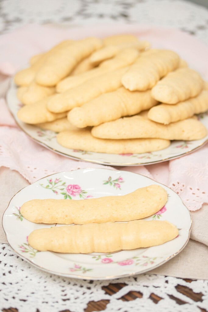 Two floral china plates of baked sourdough ladyfingers on a pink linen a large plate piled high and a small plate with two individual ladyfingers on a lace doily