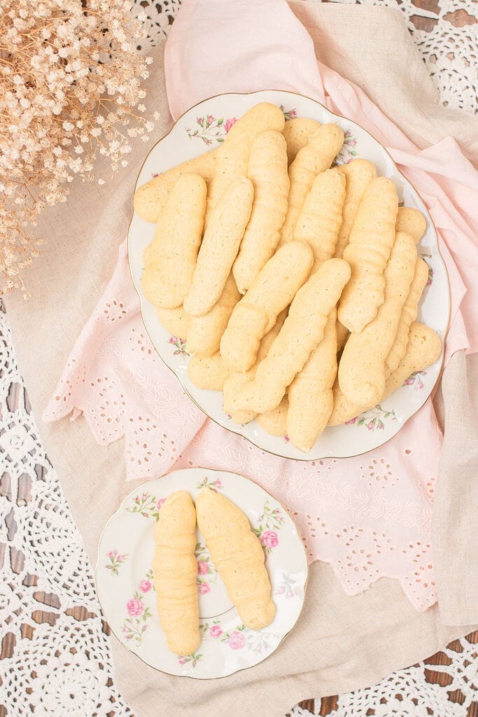 Overhead flat lay of a large floral china plate piled with sourdough ladyfingers and a small plate with two ladyfingers, on a pink eyelet linen and lace doily with dried baby's breath