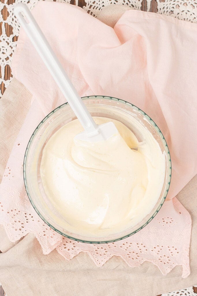 Overhead view of finished mascarpone cream folded together with a white spatula in a glass bowl, on a pink eyelet linen and lace doily