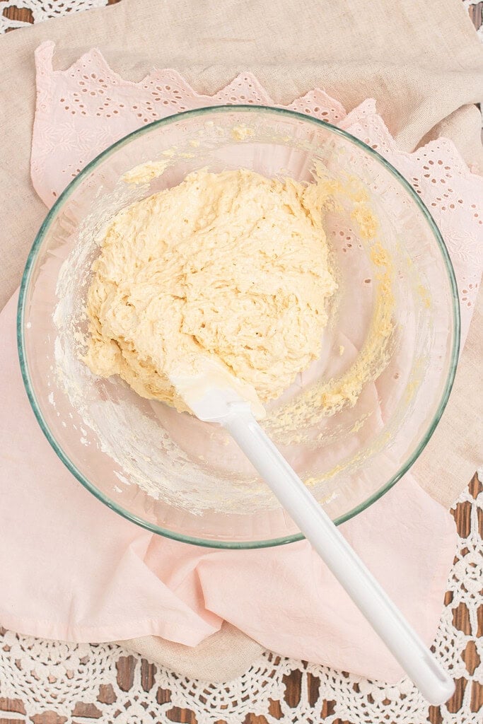 Overhead view of sourdough ladyfinger batter in a glass mixing bowl with a white spatula, on a pink linen and lace doily