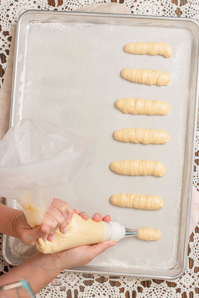 Hands piping sourdough ladyfinger batter from a pastry bag onto a parchment-lined silver baking sheet, with several piped ladyfingers already in a row
