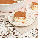 Slice of tiramisu with sourdough ladyfingers on a floral china plate with a silver fork, with the full tiramisu dish and a second serving plate in the soft background on a pink linen