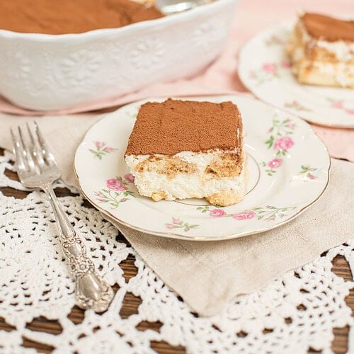 Slice of tiramisu with sourdough ladyfingers on a floral china plate with a silver fork, with the full tiramisu dish and a second serving plate in the soft background on a pink linen