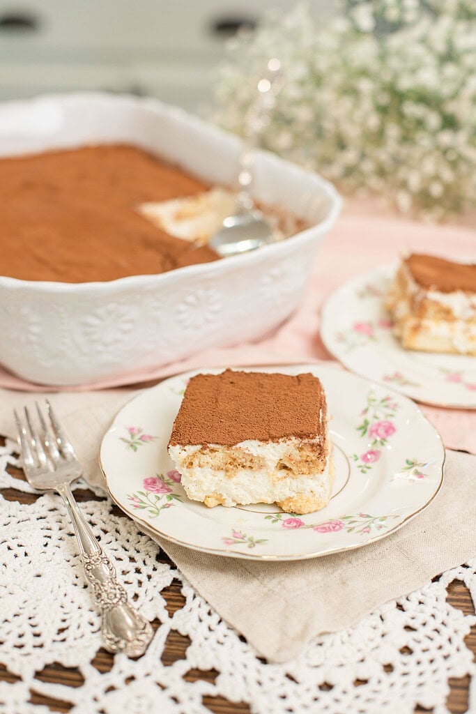 Slice of tiramisu with sourdough ladyfingers on a floral china plate with a silver fork, with the full tiramisu dish and a second serving plate in the soft background on a pink linen