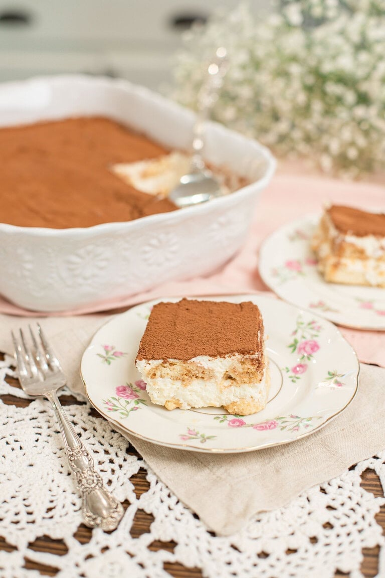 Slice of tiramisu with sourdough ladyfingers on a floral china plate with a silver fork, with the full tiramisu dish and a second serving plate in the soft background on a pink linen