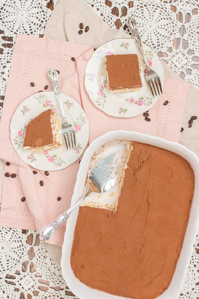 Overhead flat lay of a white baking dish of tiramisu with one corner removed, two slices served on floral china plates with silver forks, scattered coffee beans on a pink linen and lace doily