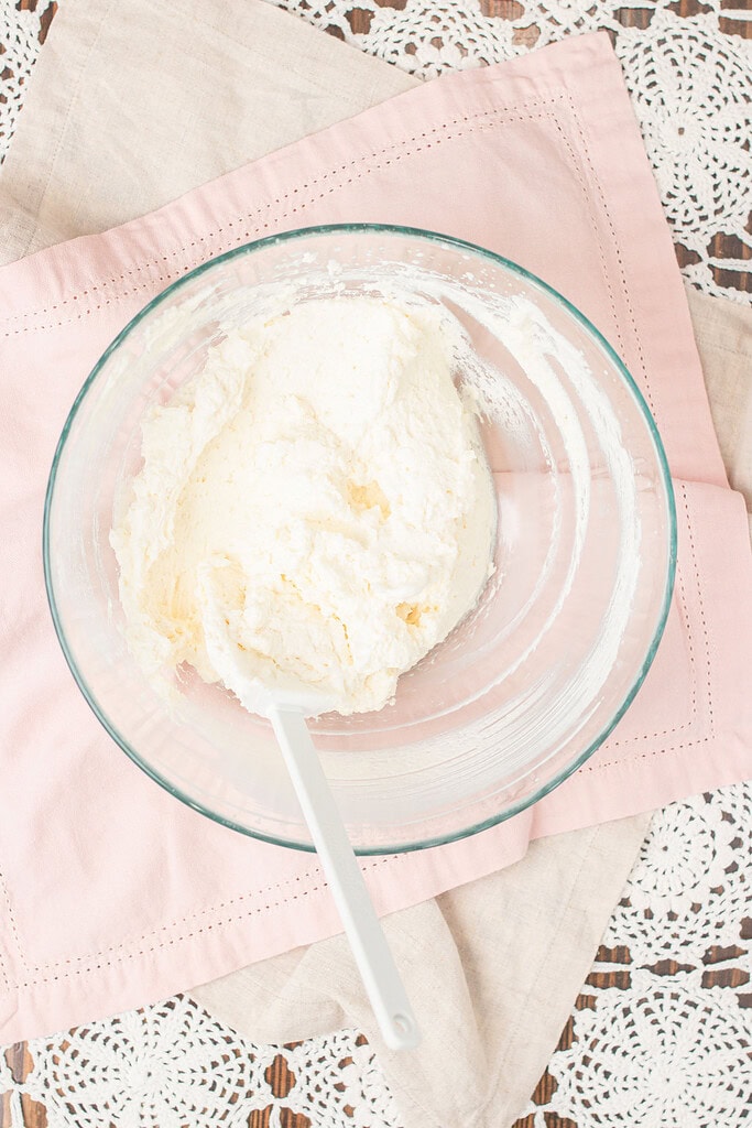 Overhead view of whipped mascarpone cream filling in a glass bowl with a white spatula, on a pink linen and lace doily