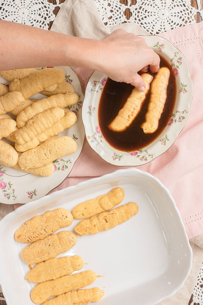 Hand dipping sourdough ladyfingers into a floral china plate of coffee, with coffee-soaked ladyfingers being arranged in a white baking dish and a plate of dry ladyfingers on the left
