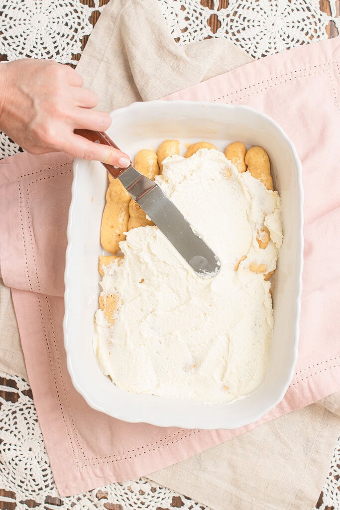 Hand spreading mascarpone cream over a layer of sourdough ladyfingers in a white baking dish with an offset spatula, on a pink linen and lace doily