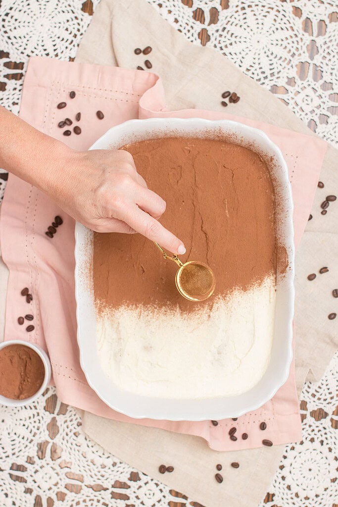 Hand dusting cocoa powder over a white baking dish of tiramisu using a gold mesh sieve, with a small bowl of cocoa powder and scattered coffee beans on a pink linen
