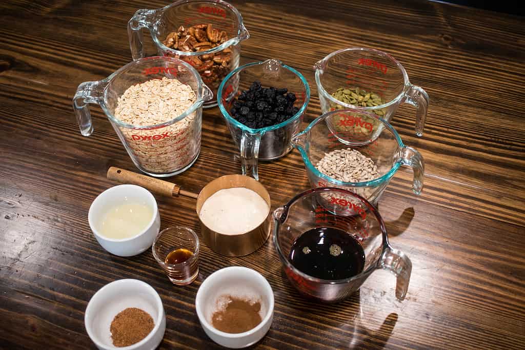Ingredients for making sourdough granola in measuring cups, bowls, on a wooden counter top in a kitchen.