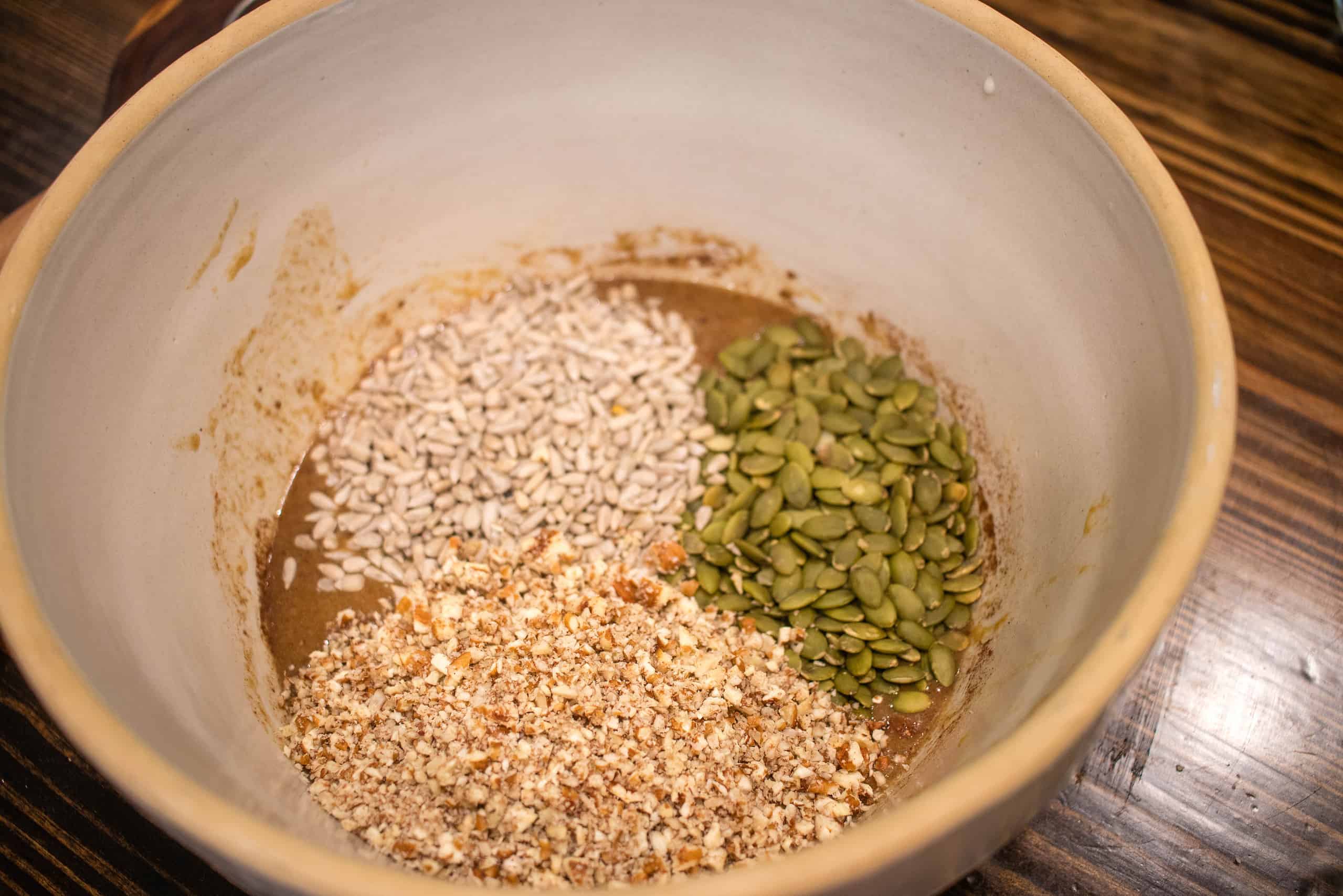 A large ceramic mixing bowl on a wooden counter top in a cottage kitchen has nuts and seeds in a sourdough mixture.