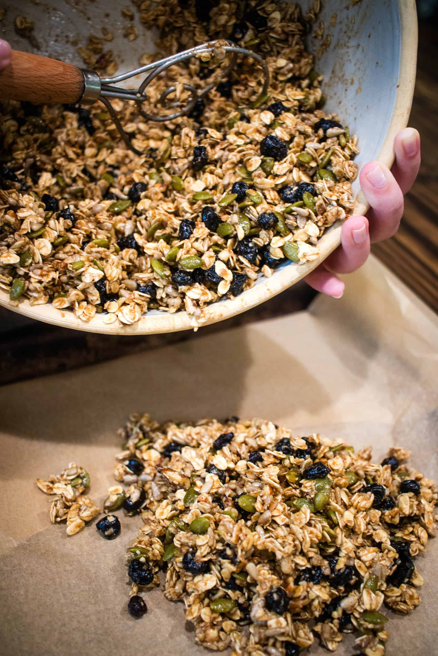 A women is pouring a uncooked granola mixture onto a baking pan lined with parchment paper in a cottage kitchen.