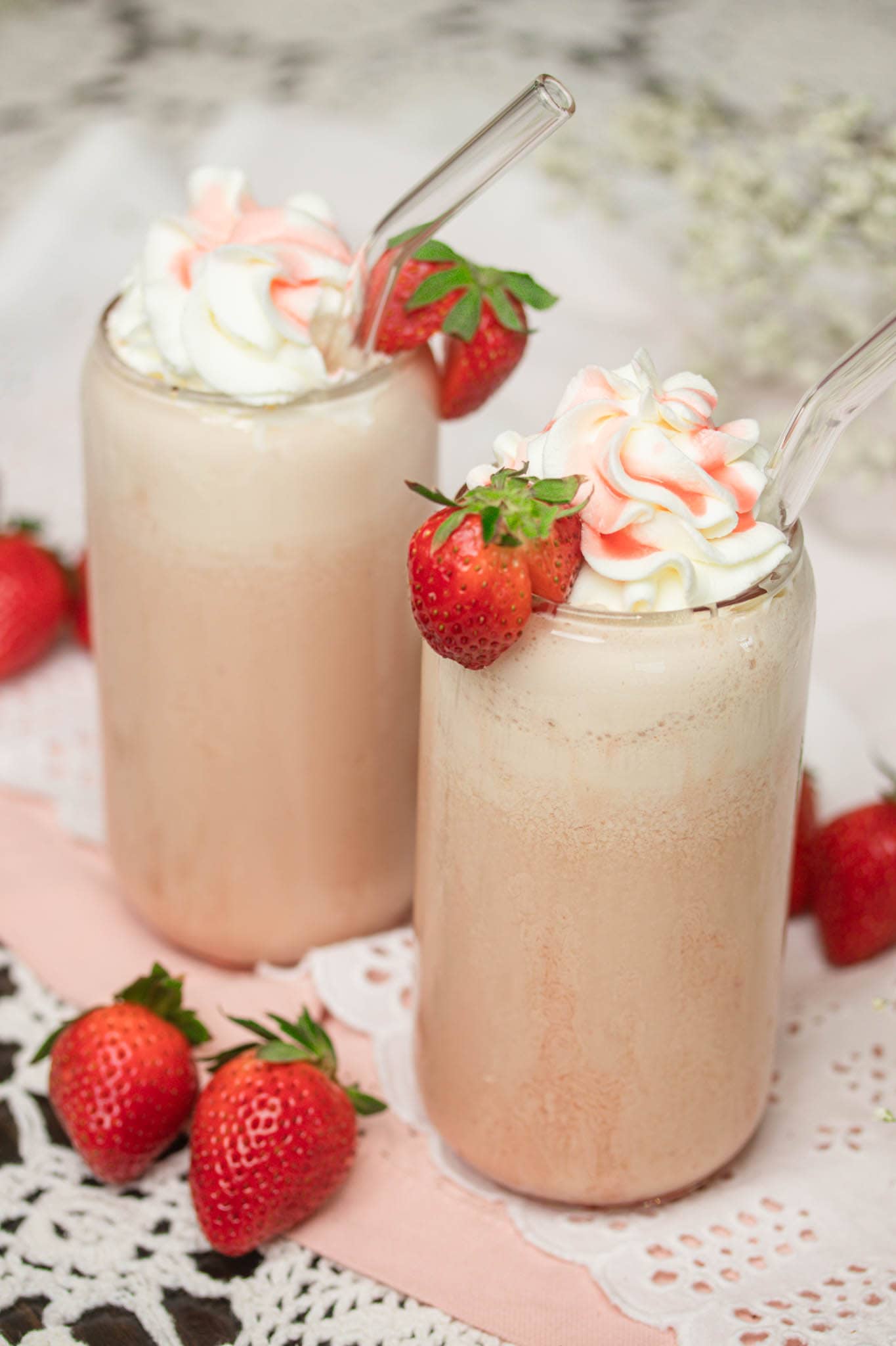 Two tall strawberry frappes in glass jars topped with whipped cream and fresh strawberries, served with clear straws on a lace-covered table.