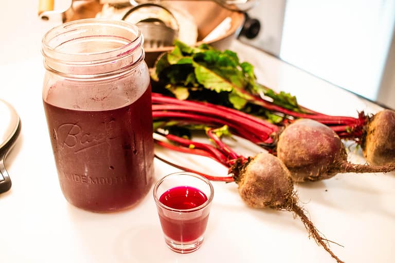 A mason jar and a shot glass or beet kvass and three beets on a marble slab in a kitchen.