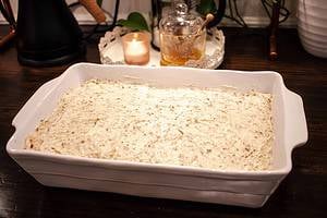 A carrot cake with cream cheese frosting in a 9x13 white baking dish on a wooden counter top in a cottage kitchen with candles and a coffee press behind the cake dish.