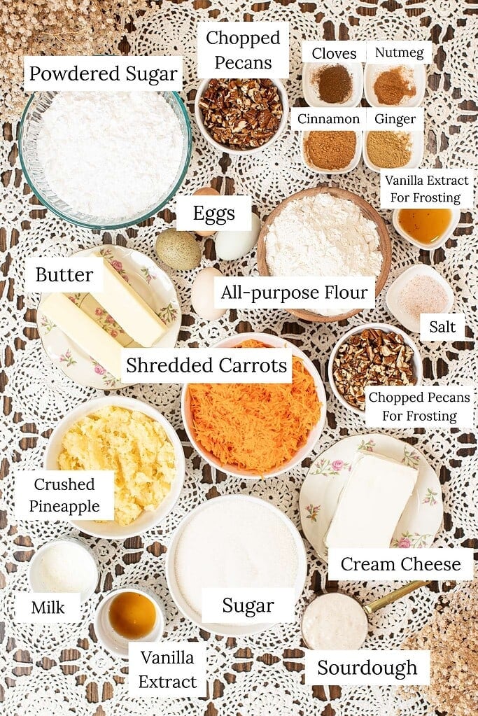 Ingredients for sourdough carrot cake and cream cheese frosting arranged on a table, including flour, sugar, sourdough starter, shredded carrots, crushed pineapple, butter, eggs, chopped pecans, cream cheese, powdered sugar, milk, vanilla extract, cinnamon, ginger, cloves, nutmeg, and salt.