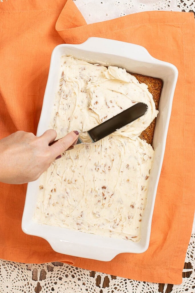 Offset spatula spreading cream cheese frosting over cooled carrot cake in a white baking dish.