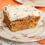 Close-up of a slice of carrot cake with thick cream cheese frosting on a floral plate, showing a moist crumb with shredded carrots and nuts.