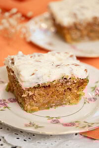 Close-up of a slice of carrot cake with thick cream cheese frosting on a floral plate, showing a moist crumb with shredded carrots and nuts.