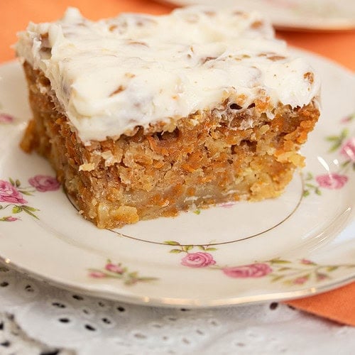 Close-up of a slice of carrot cake with thick cream cheese frosting on a floral plate, showing a moist crumb with shredded carrots and nuts.