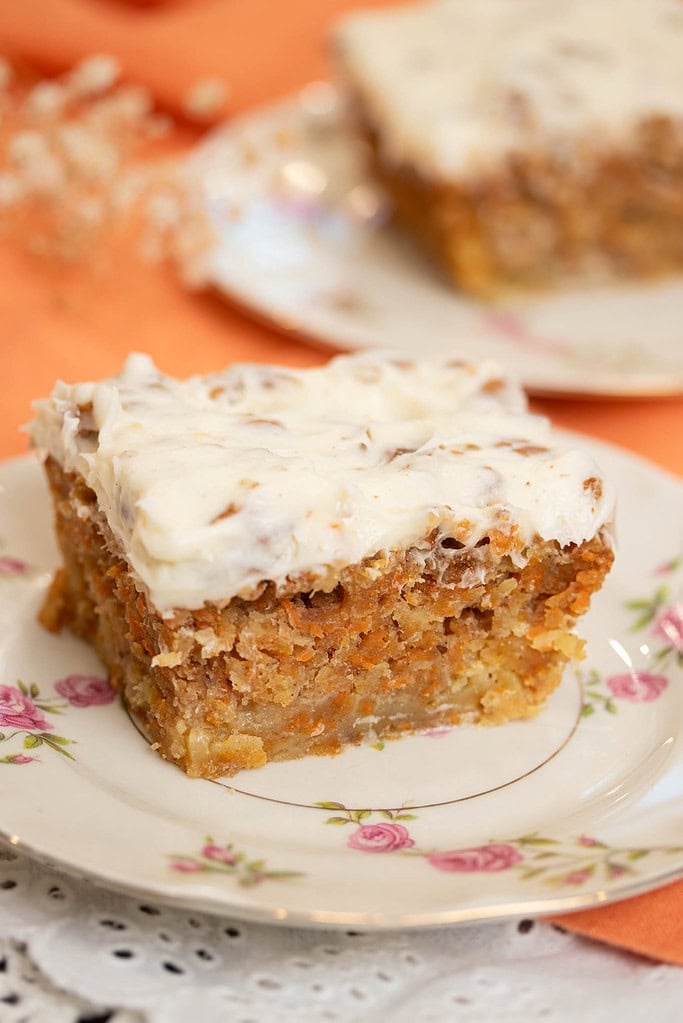 Close-up of a slice of carrot cake with thick cream cheese frosting on a floral plate, showing a moist crumb with shredded carrots and nuts.