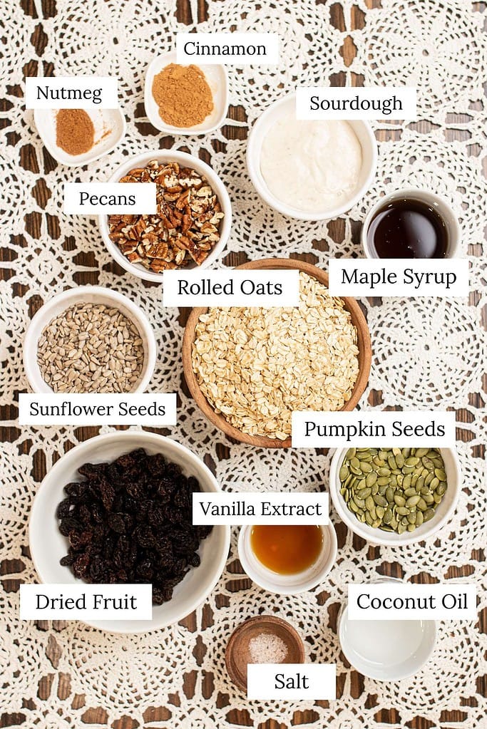 Ingredients for sourdough granola arranged in small bowls, including rolled oats, sourdough starter, maple syrup, coconut oil, pecans, seeds, dried fruit, cinnamon, nutmeg, vanilla extract, and salt.