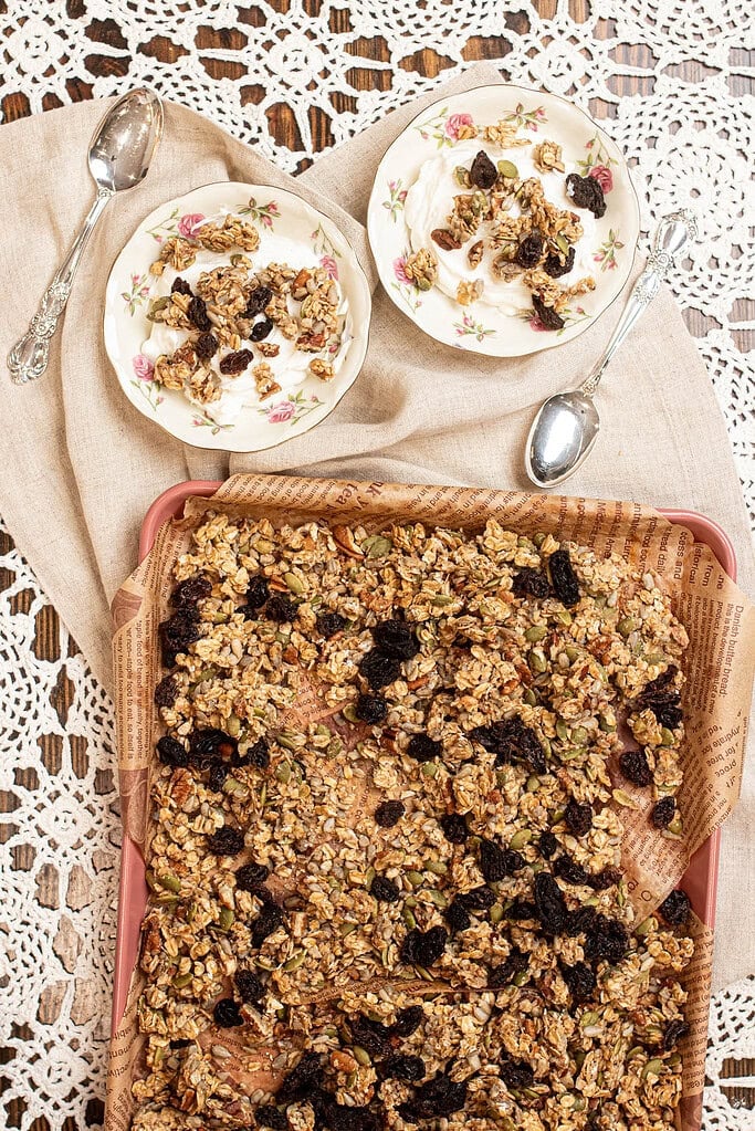 Sourdough granola served over creamy yogurt in floral dishes with silver spoons, showing crisp clusters and dried fruit.