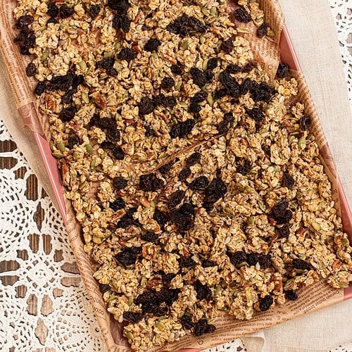 Sourdough granola spread on a parchment-lined baking sheet, filled with rolled oats, pecans, pumpkin seeds, sunflower seeds, and raisins baked into golden clusters.