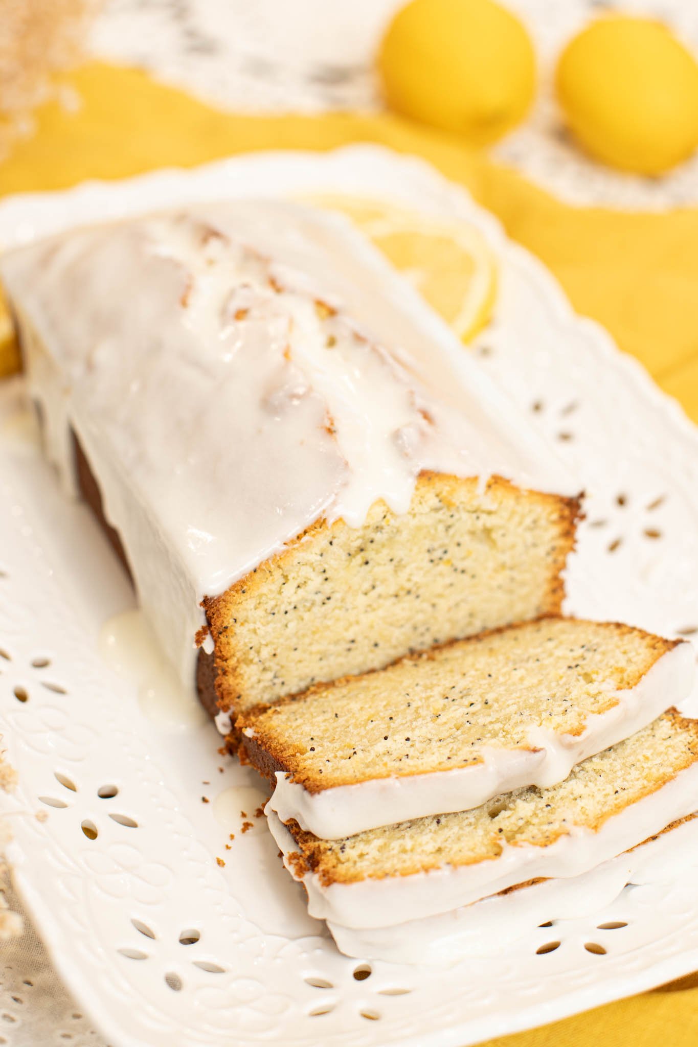 Sliced sourdough lemon poppy seed pound cake with thick lemon glaze on a white serving platter, fresh lemons in the background.
