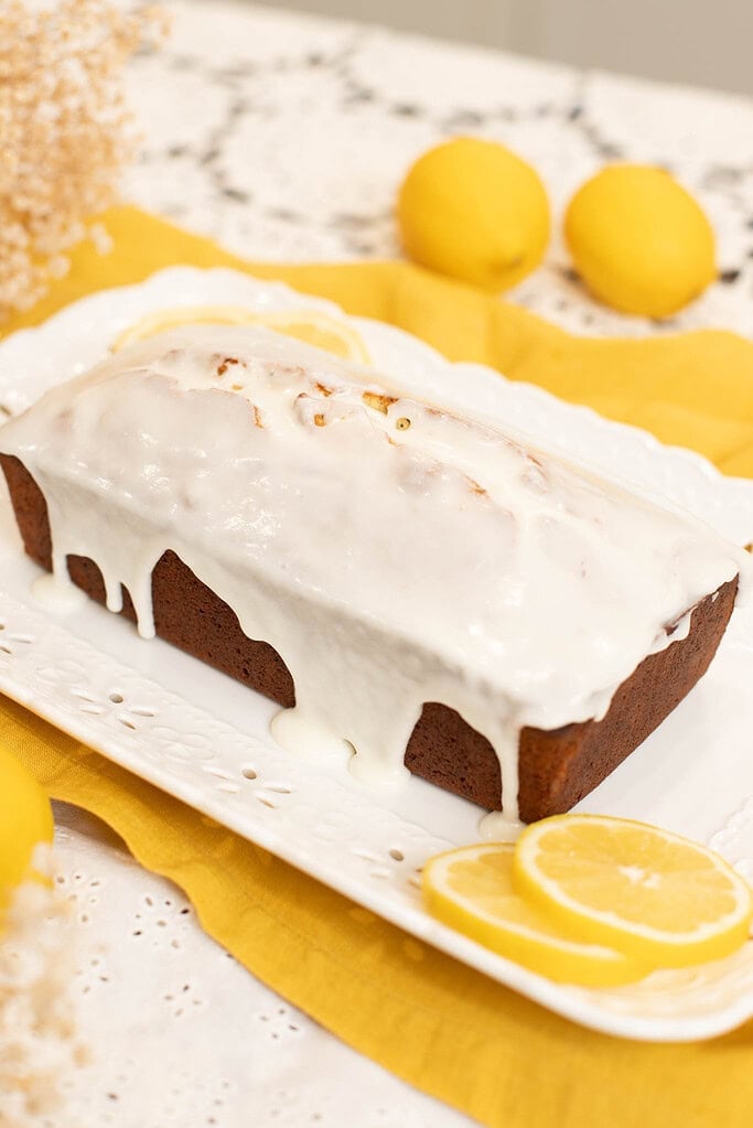 Whole sourdough lemon poppy seed pound cake topped with thick lemon glaze on a white platter with fresh lemon slices and yellow linen.