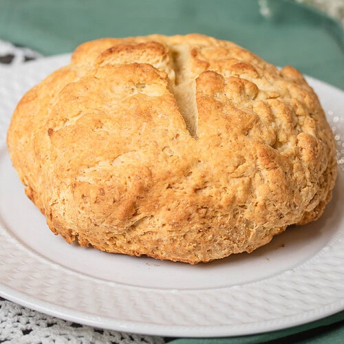 Freshly baked traditional Irish soda bread with a golden crust and a deep cross cut on top, sitting on a white plate with a green linen napkin on a lace tablecloth.
