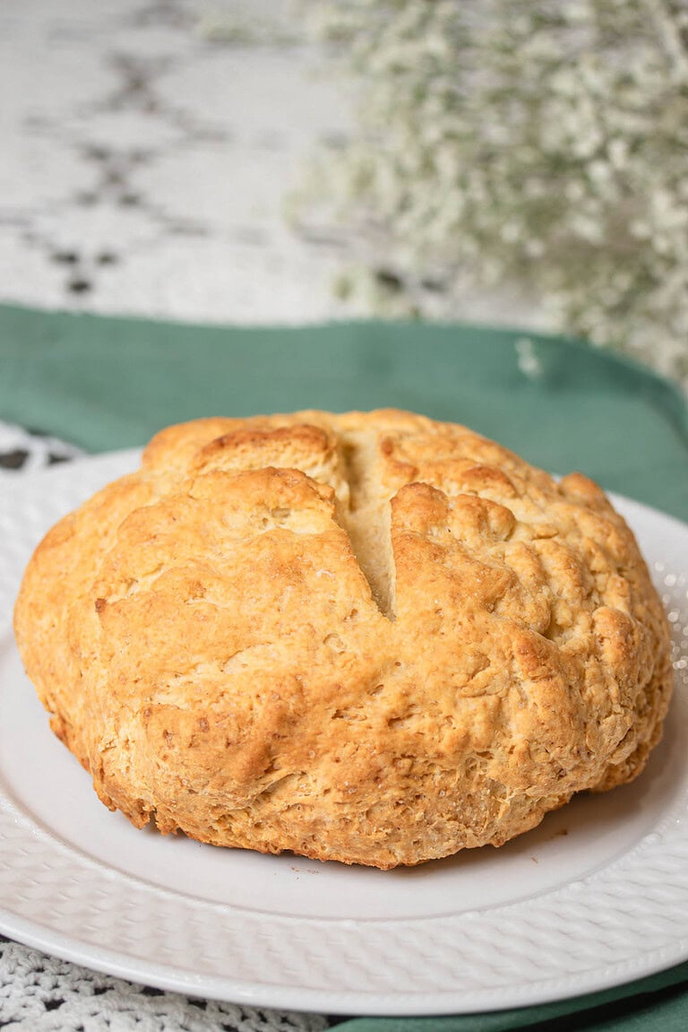 Freshly baked traditional Irish soda bread with a golden crust and a deep cross cut on top, sitting on a white plate with a green linen napkin on a lace tablecloth.