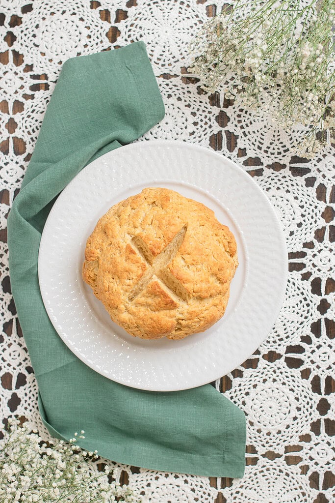 Overhead view of a round loaf of Irish soda bread with a cross cut into the top, resting on a white plate with a green cloth napkin on a lace tablecloth.