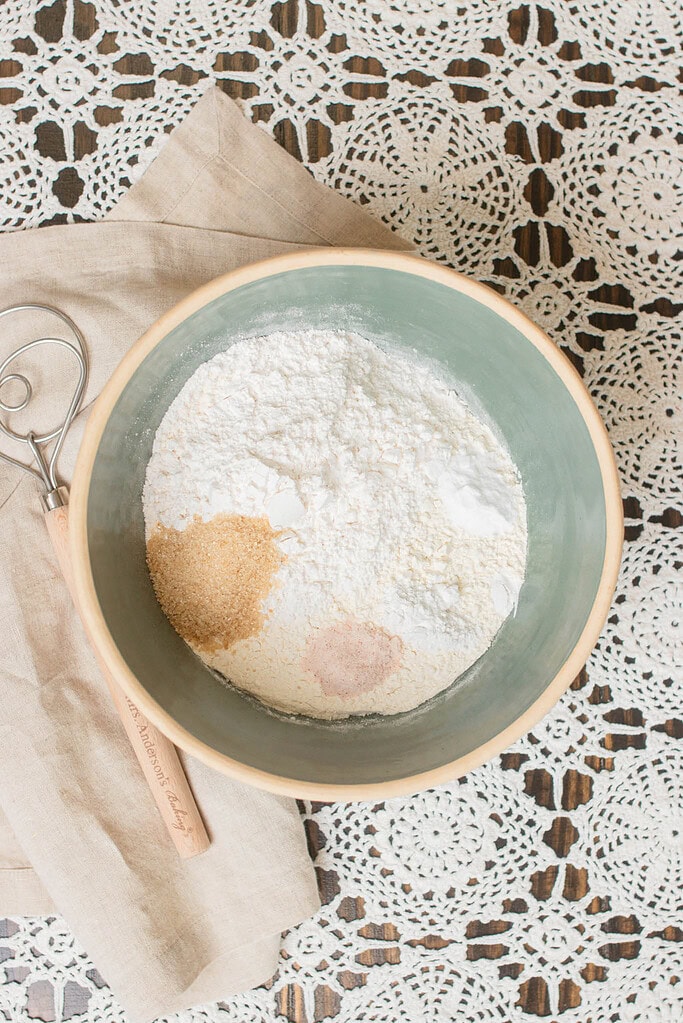 Dry ingredients for Irish soda bread in a mixing bowl, including flour, sugar, salt, baking soda, and cream of tartar, with a dough whisk resting beside the bowl.